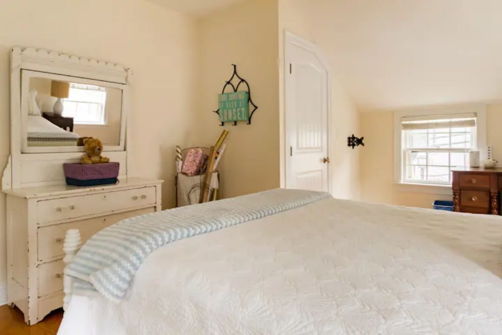 A bedroom with a large bed, a dresser with a mirror, a small table with a teddy bear, a wall-mounted sign, and a window.