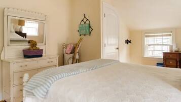 A bedroom with a large bed, a dresser with a mirror, a small table with a teddy bear, a wall-mounted sign, and a window.