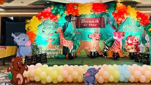 Un escenari decorat amb globus de colors i dibuixos d'animals de cartó animat, com ara elefants, girafes i tigres, amb un cartell que diu 'Happy Birthday'.