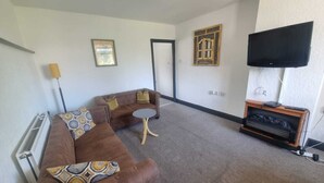 A living room with a brown sofa, a small round table, a flat-screen TV on a wooden stand, and two framed pictures on the wall. - Windsor Apartments (Dartford)