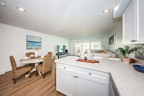 A modern kitchen with white cabinets, a dining area with wicker chairs, and a living room with a sofa and a television.