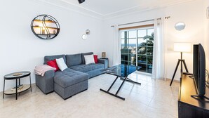 A modern living room with a grey sectional sofa, a glass coffee table, a round wall clock, and a large window with a view of palm trees. - Superbe Appartement 2 Chambres Avec vue mer (Albufeira)