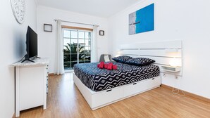 A modern bedroom with a bed, a desk, a TV, and a window with a view of greenery. - Superbe Appartement 2 Chambres Avec vue mer (Albufeira)