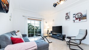 A modern living room with a grey sofa, a red pillow, a pink throw blanket, a black TV stand, and a black chair. - Superbe Appartement 2 Chambres Avec vue mer (Albufeira)