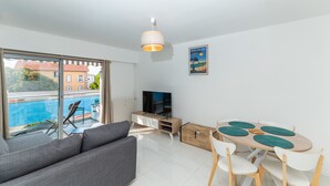 A modern living room with a sofa, TV, dining table with chairs, and a view of a swimming pool. - (012) L'armenonville à la plage (Antibes)