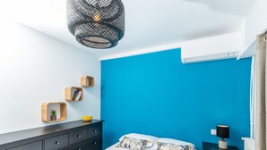 A bedroom with a bed, a black dresser, a small table, a hanging light fixture, and a blue accent wall. - (012) L'armenonville à la plage (Antibes)