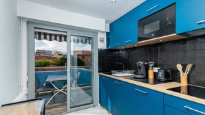 A modern kitchen with blue cabinets, a black tiled backsplash, and a sliding glass door leading to a balcony with outdoor furniture. - (012) L'armenonville à la plage (Antibes)