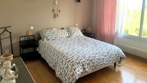 Ein Schlafzimmer mit einem Bett, zwei Nachttischen, einem Wandbehang im Traumfänger-Stil, einem Fenster mit Vorhängen und einem Heizkörper.