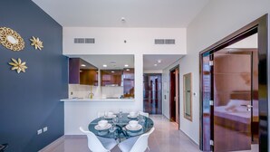 A modern hotel room with a dining area featuring a glass table set for six, white chairs, and a kitchen in the background. - Tanin - Unique Modern Apt with Stunning Canal View (Dubai)