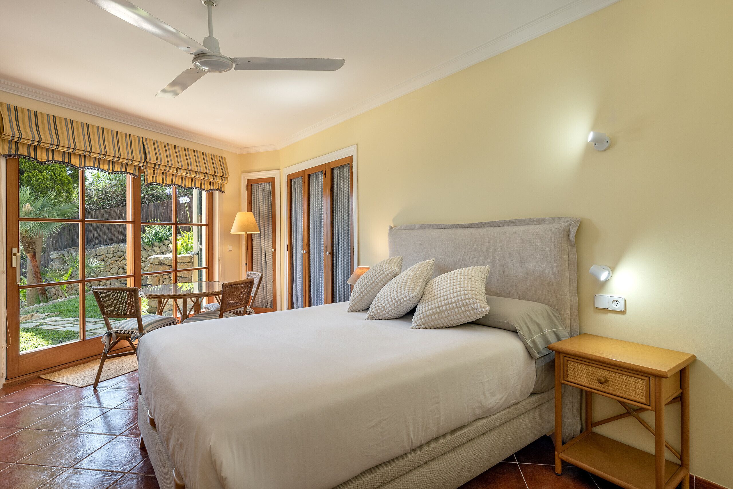Un dormitorio con cama, ventilador de techo, mesita de noche y ventana con cortinas.