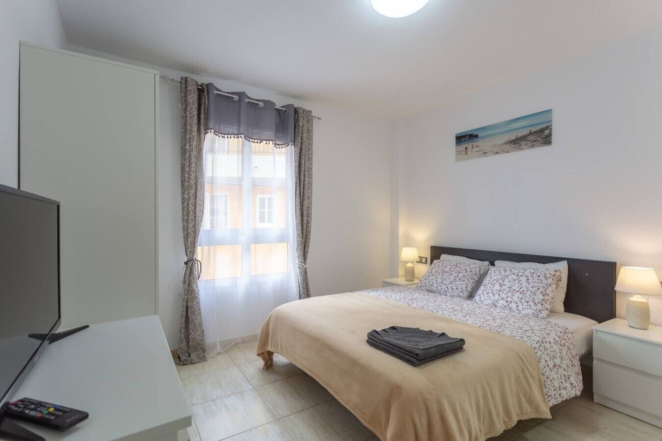 Un dormitorio con cama, lámparas de noche, televisión y ventana con cortinas.