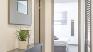 A hallway with a mirror, a potted plant, a console table, and a door leading to a bedroom with a bed and a window.