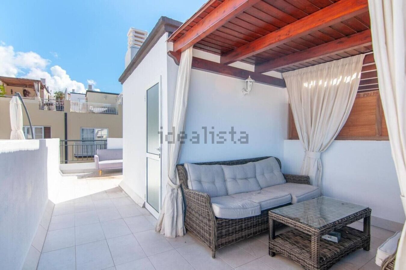 Un balcon avec un canapé en osier et une petite table, un mur blanc agrémenté d’un luminaire et un plafond en bois.