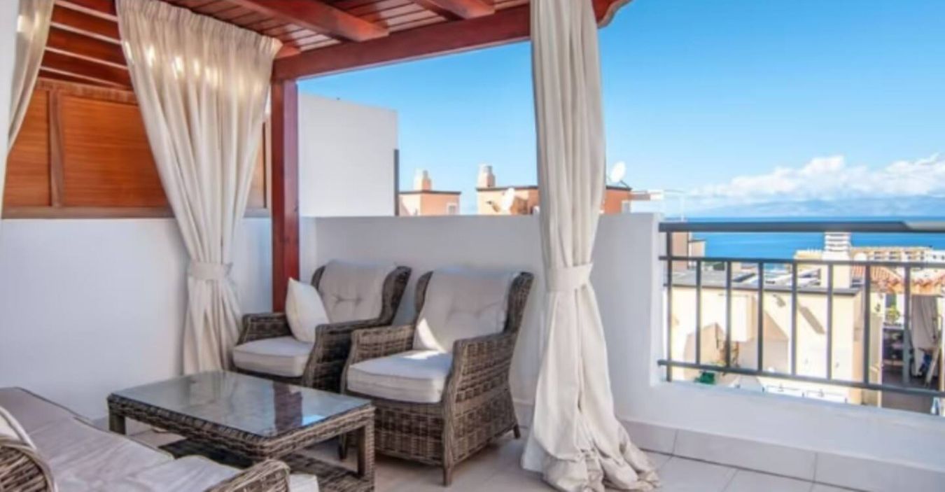 A balcony with wicker furniture and white curtains, overlooking a cityscape.