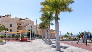 Un parque infantil con palmeras, una zona de juegos roja y edificios al fondo.