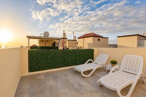 Eine Dachterrasse mit zwei weißen Liegestühlen, einem grünen Hecken und Blick auf Gebäude im Hintergrund.