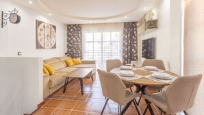 Un comedor con una mesa redonda para cuatro personas, un sofá con cojines amarillos y un reloj en la pared.