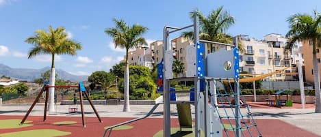 Un parque infantil con columpios y estructuras para escalar, rodeado de palmeras y edificios.
