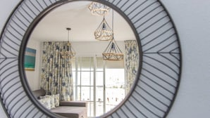 Un miroir rond doté d’un cadre géométrique, qui reflète une pièce avec un rideau à motifs, un luminaire suspendu et une fenêtre avec des stores.