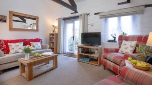 A cozy living room with a sofa, a coffee table, a TV on a wooden stand, and a bookshelf. - Rose Cottage in Marldon, Paignton, Devon (Paignton)