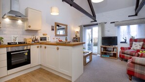 A modern kitchen with white cabinets, a built-in oven, and a microwave. There is a wooden dining table with a TV and a red plaid sofa in the living area. - Rose Cottage in Marldon, Paignton, Devon (Paignton)