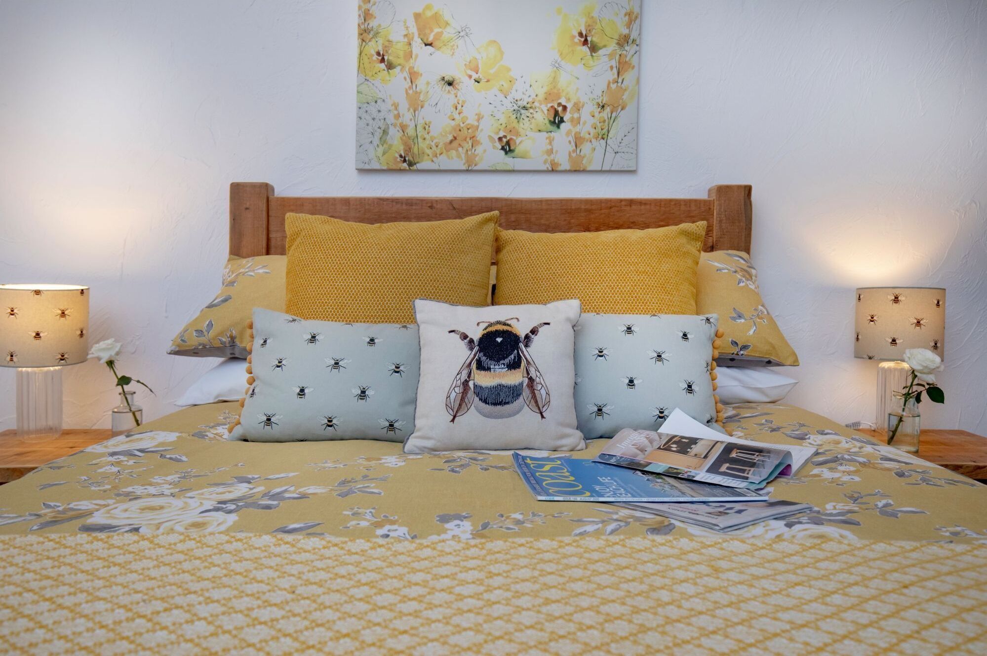 Een bed met een bloemenbedekking, meerdere kussens met bijenmotieven, en een tijdschrift op het bed.