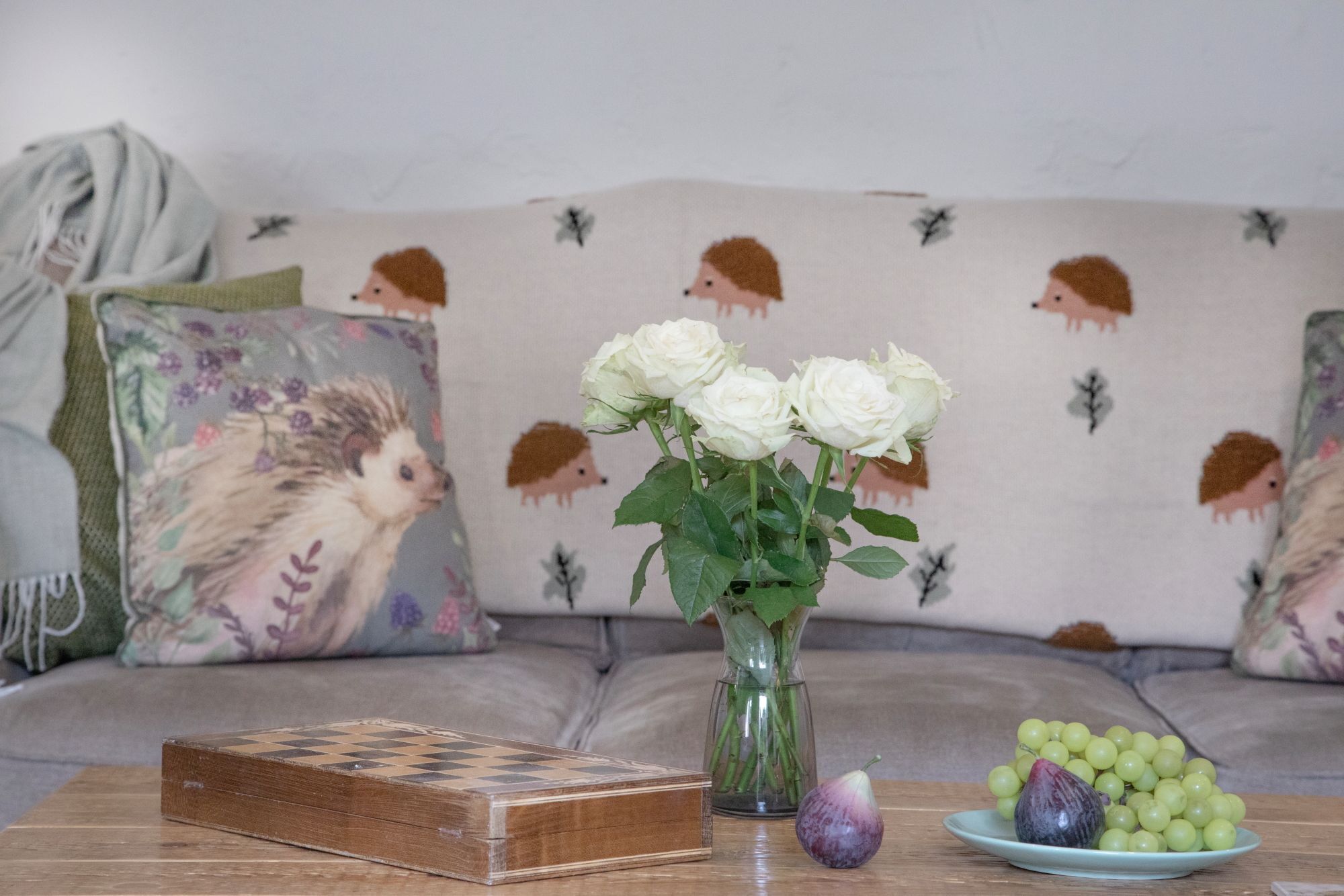 A living room with a sofa, a vase with white roses, a chessboard, and a plate of grapes.