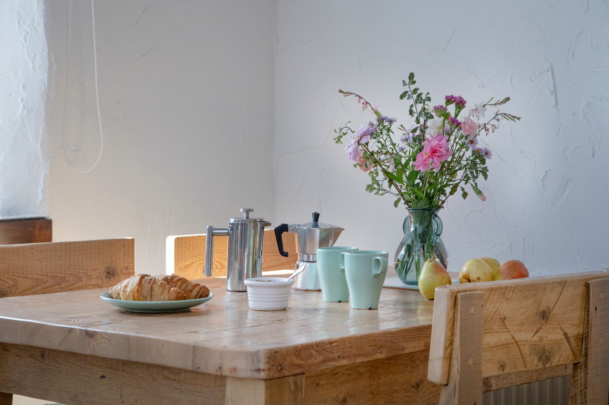 Una mesa de madera con un croissant, una cafetera, un florero con flores y algunas manzanas.