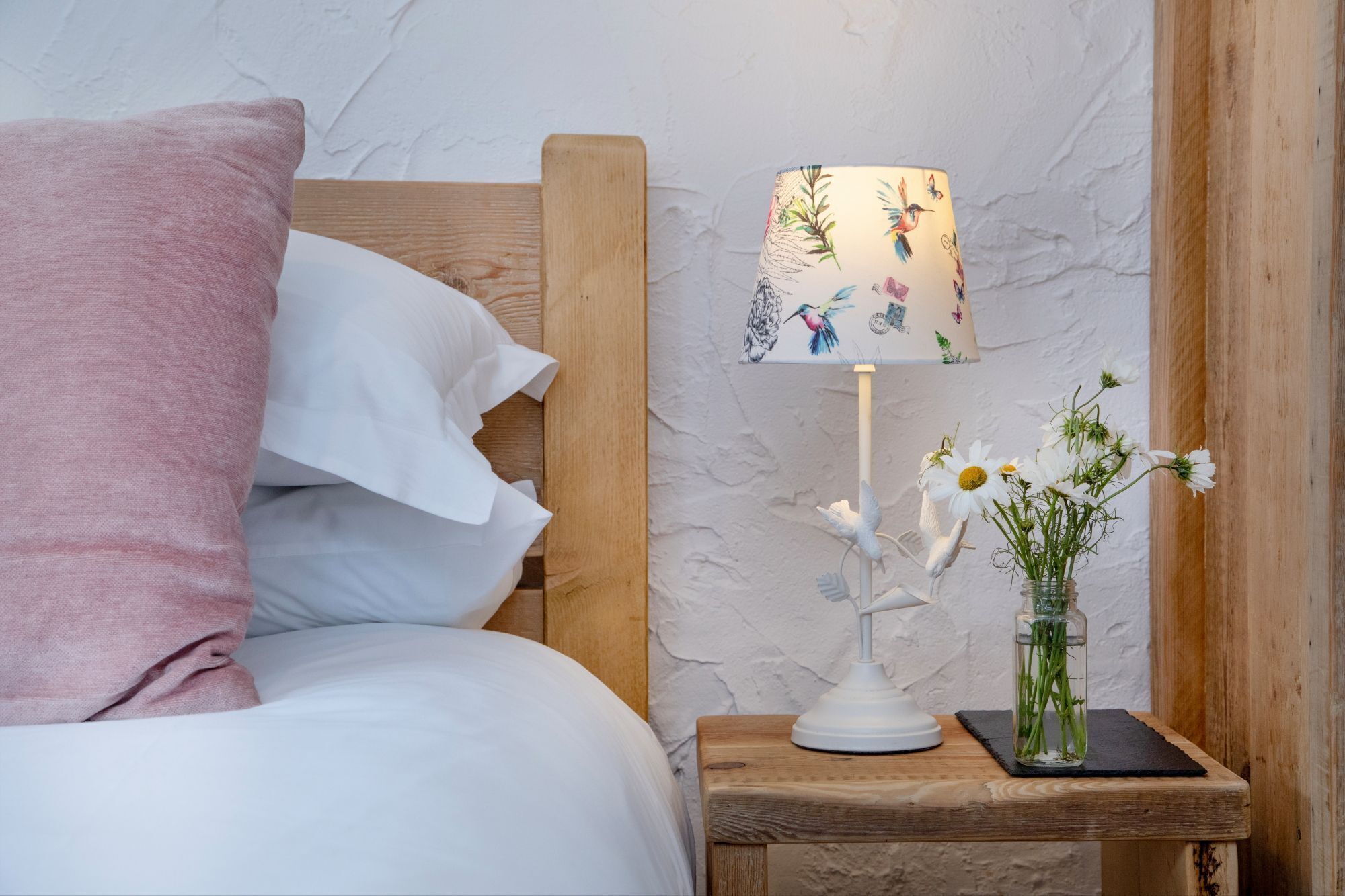 Ein ordentlich bezogenes Bett mit einem rosafarbenen Kopfkissen und weißen Textilien, ein Nachttisch aus Holz mit einer weißen, floral verzierten Lampe und eine Vase mit weißen Blumen.