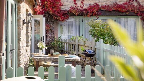 Un espace extérieur rustique aménagé avec une table en bois, un barbecue et une clôture blanche en piquets.