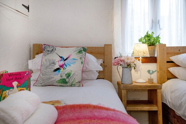 A neatly made bed with a colorful pillow, a bedside table with a lamp and flowers, and a window with sheer curtains. - Daisy Cottage - Sleeps 4 guests in 2 bedrooms (Paignton)