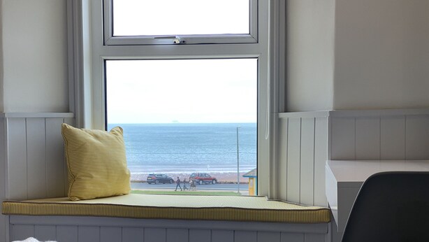 Una stanza con vista sull'oceano attraverso una finestra, una panca con un cuscino giallo, una sedia e un letto con asciugamani piegati.