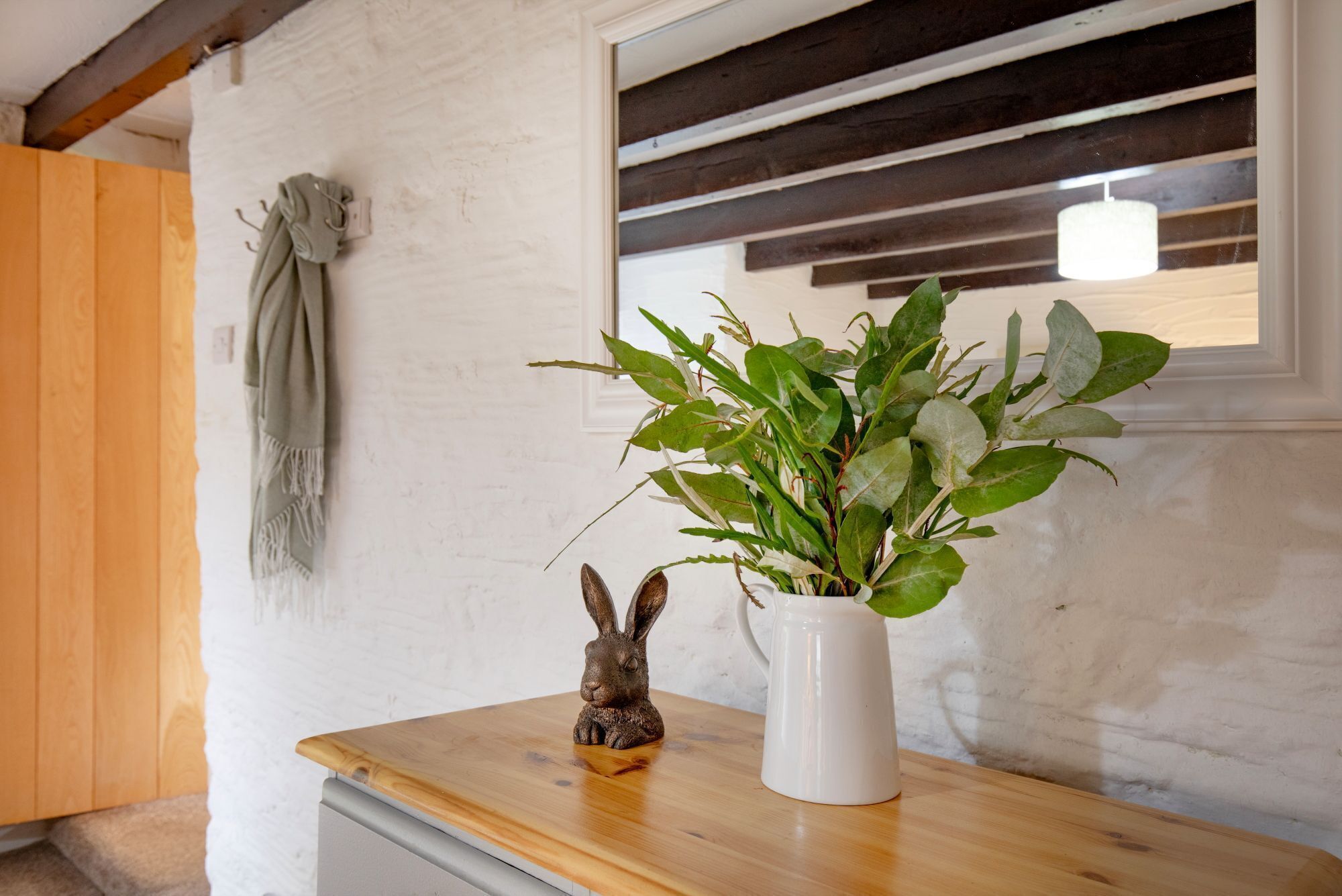 Una mesa de madera con un jarrón blanco que contiene plantas verdes y una pequeña figurina de conejo.