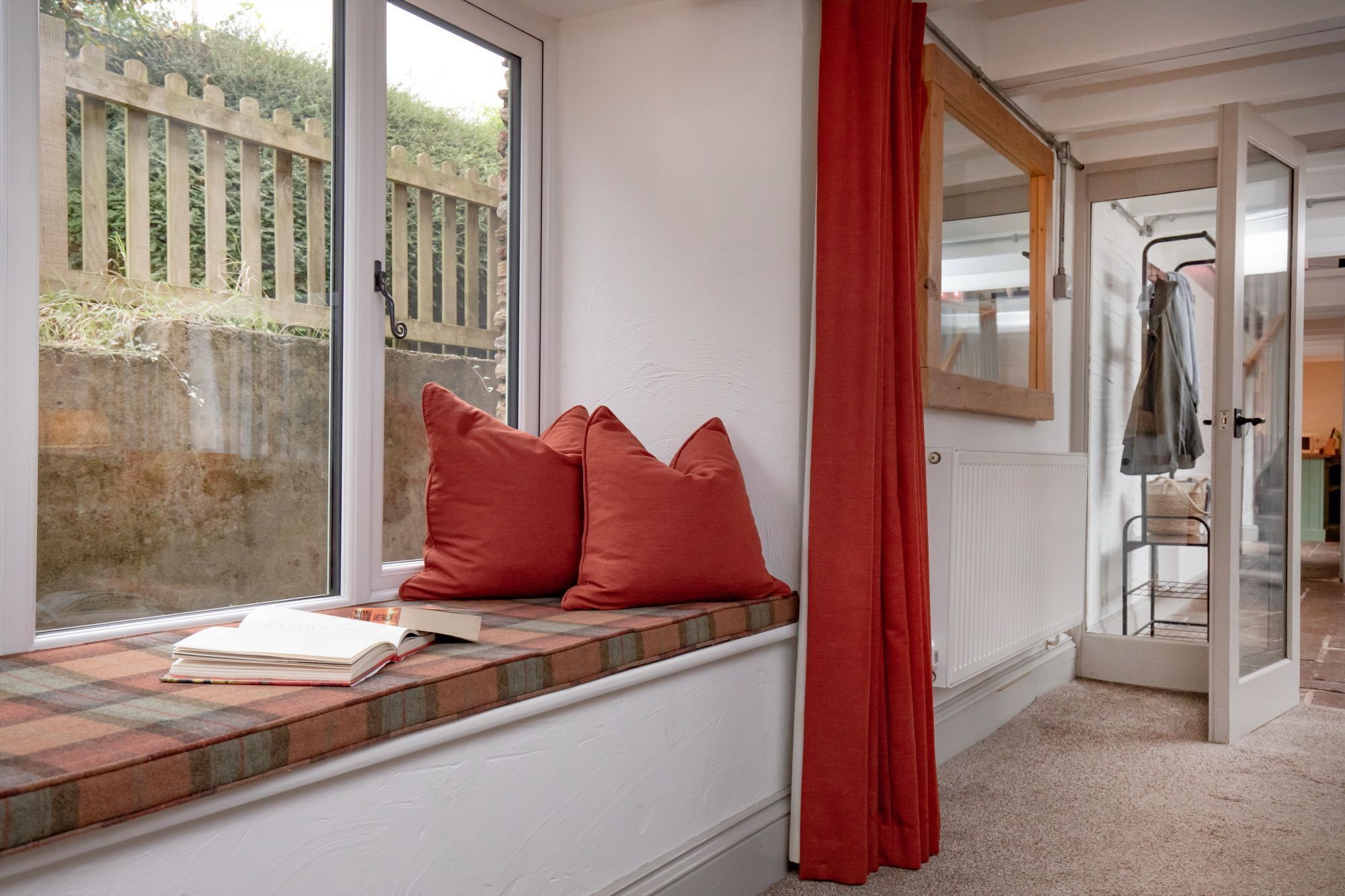 Una habitación con asiento junto a la ventana, un radiador y una puerta que da a otra estancia.