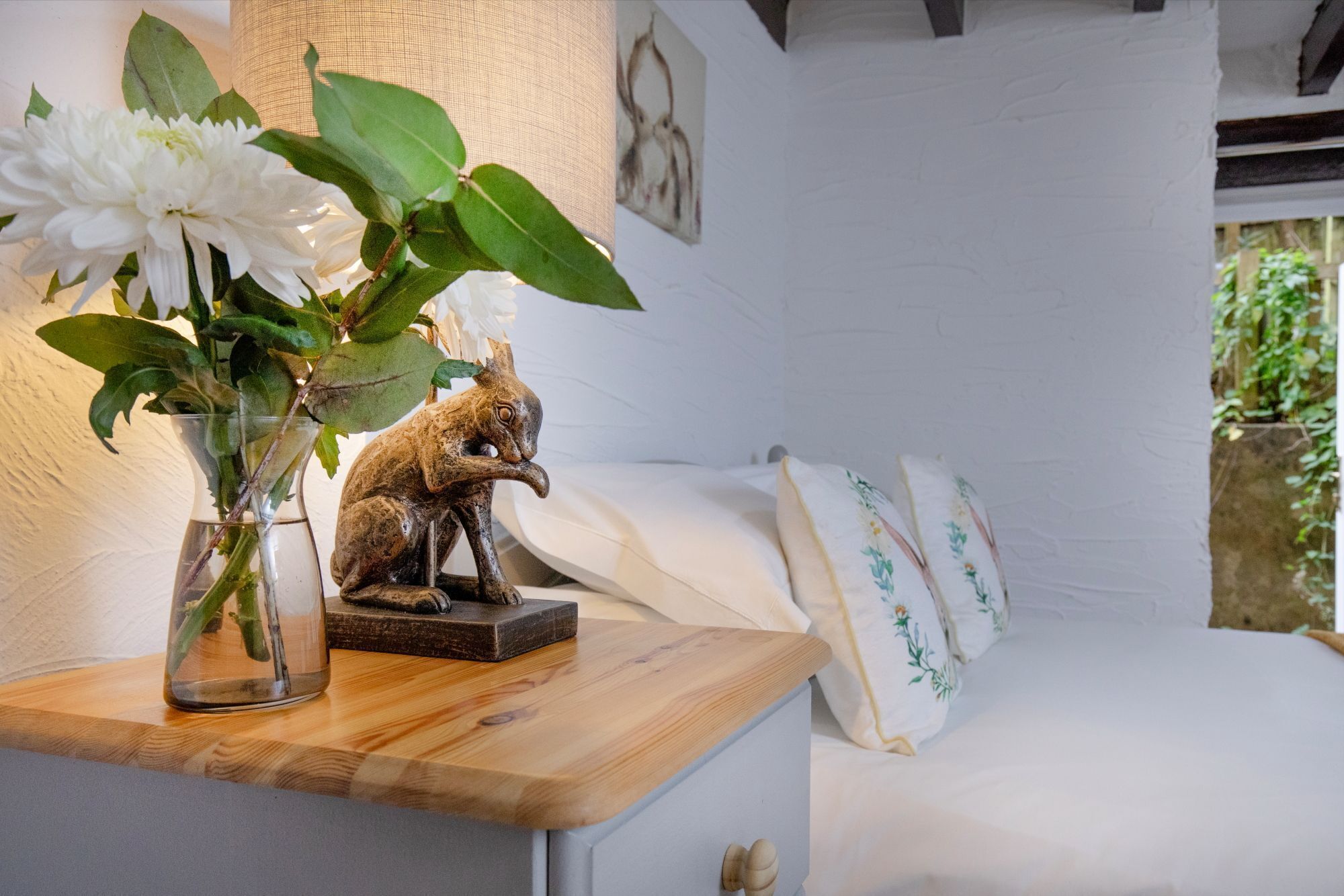 A bedroom with a wooden bedside table, a vase with white flowers, a small animal figurine, and a bed with white bedding and floral pillows.