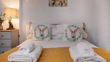 A neatly made bed with two pillows featuring embroidered rabbits and floral designs, a bedside table with a lamp and a vase of flowers, and a painting on the wall.