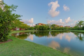 A serene lakeside view with a well-maintained lawn, a calm water body reflecting the sky, and several buildings with red roofs in the background.