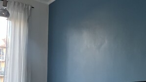 Un dormitorio con una cama frente a una pared azul, una ventana con cortinas blancas y una lámpara colgante.