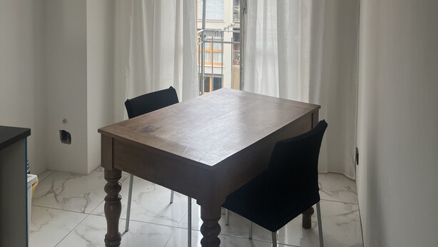 Un área de comedor con una mesa y sillas de madera, vista a un edificio contiguo a través de cortinas translúcidas, y una lámpara con forma geométrica en el techo.