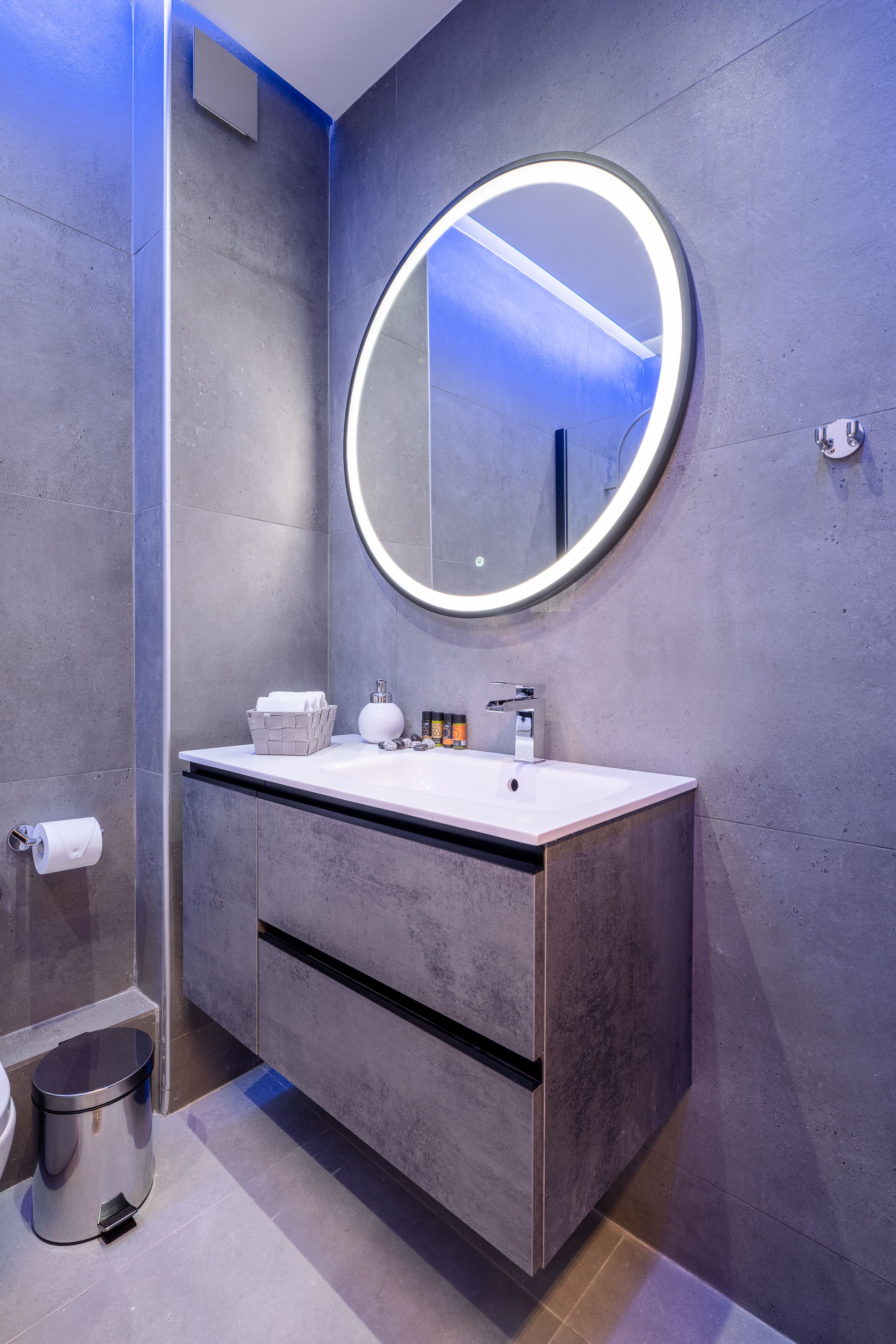 A modern bathroom with a round mirror, white sink, and grey cabinets.