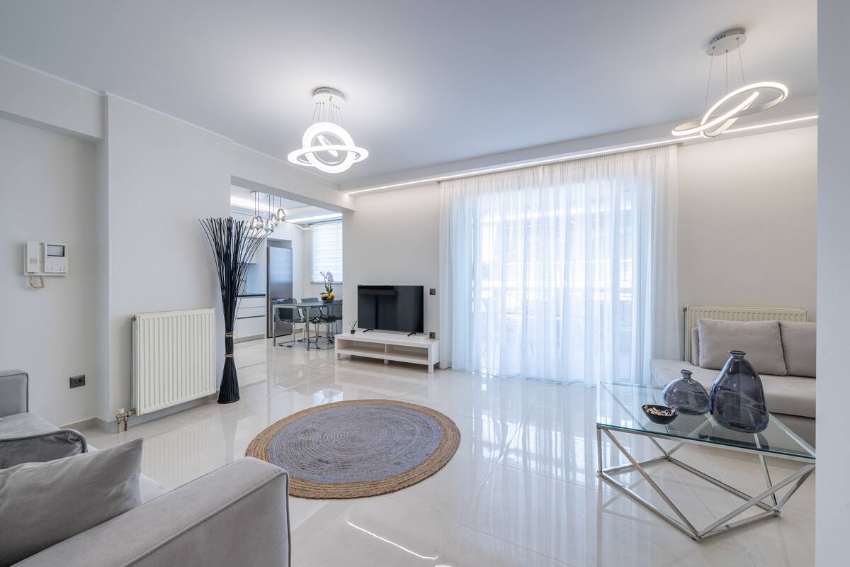 A modern living room with a glass coffee table, a sofa, a TV, and a dining area in the background.