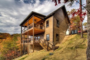 A two-story wooden cabin with a balcony on the upper floor, surrounded by a forested hillside.