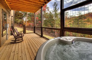 A wooden deck with a hot tub and rocking chairs, overlooking a forested area.