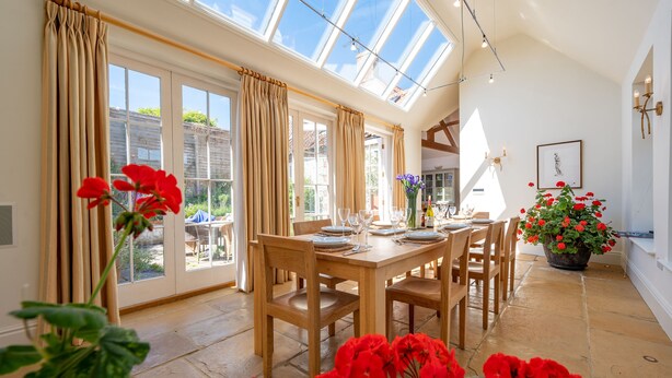 A dining area with a large table set for four, a potted plant with red flowers, and a view through the glass doors to the outside. - School House, Burnham Thorpe, Norfolk (Burnham Thorpe)