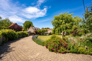 A paved driveway leading to a house with a sloped roof, surrounded by lush greenery and flowering plants.