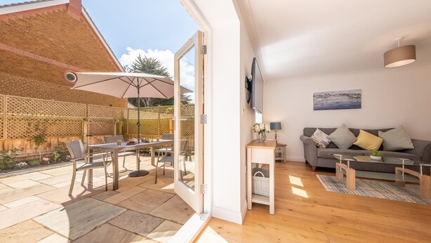 Un salon moderne avec un canapé, une table basse et un téléviseur fixé au mur. Il y a également un espace patio avec une table et des chaises, ainsi qu’une clôture en bois.