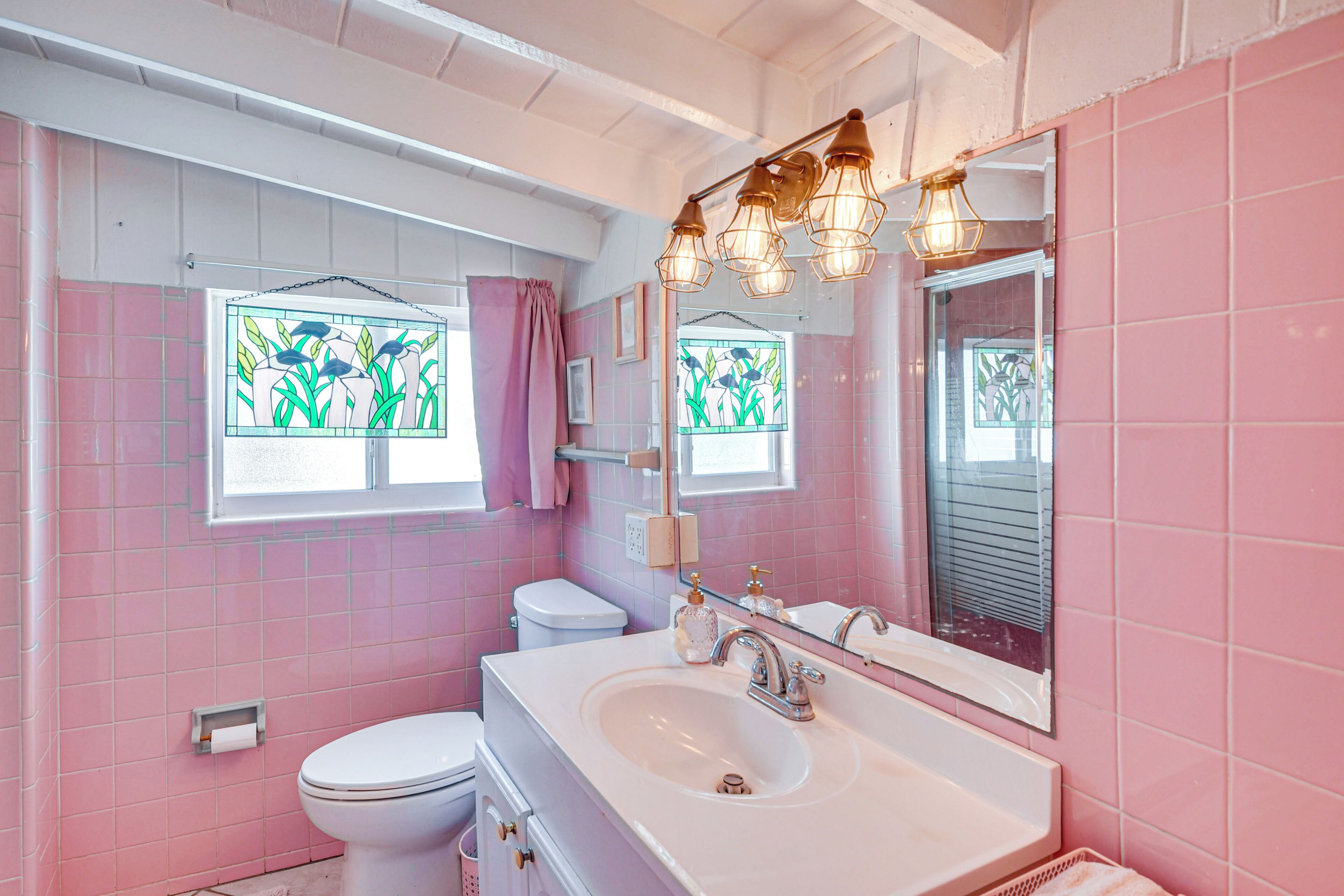 A bathroom with pink tiles, a white sink, a mirror, and a window with a view of plants.