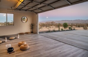 A spacious room with a large wooden floor, a yoga mat, a meditation cushion, and a view of a desert landscape.