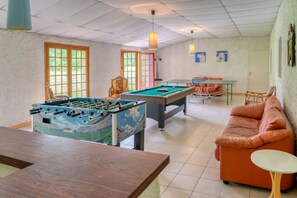 Quarto com mesa de futebol, mesa de bilhar, sofá laranja e uma pequena mesa com abajur.