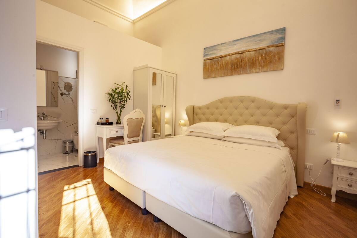 A bedroom with a large bed, a nightstand, a lamp, a mirror, a wardrobe, and a painting on the wall.